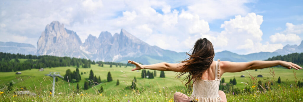 Donna di spalle che ammira il paesaggio delle Dolomiti in primavera, con prati verdi e montagne sullo sfondo sotto cielo limpido.
